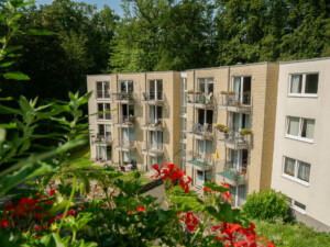 Mehrstöckiges Wohngebäude mit Balkonen und Blumenkästen, umgeben von Bäumen und Gartenanlage.