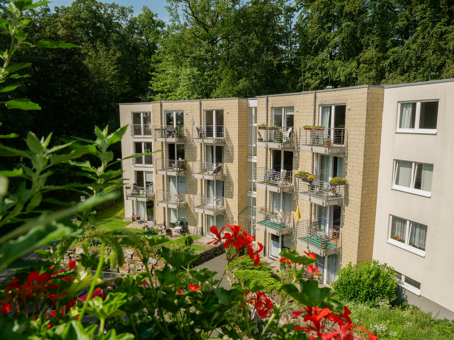 Mehrstöckiges Wohngebäude mit Balkonen und Blumenkästen, umgeben von Bäumen und Gartenanlage.