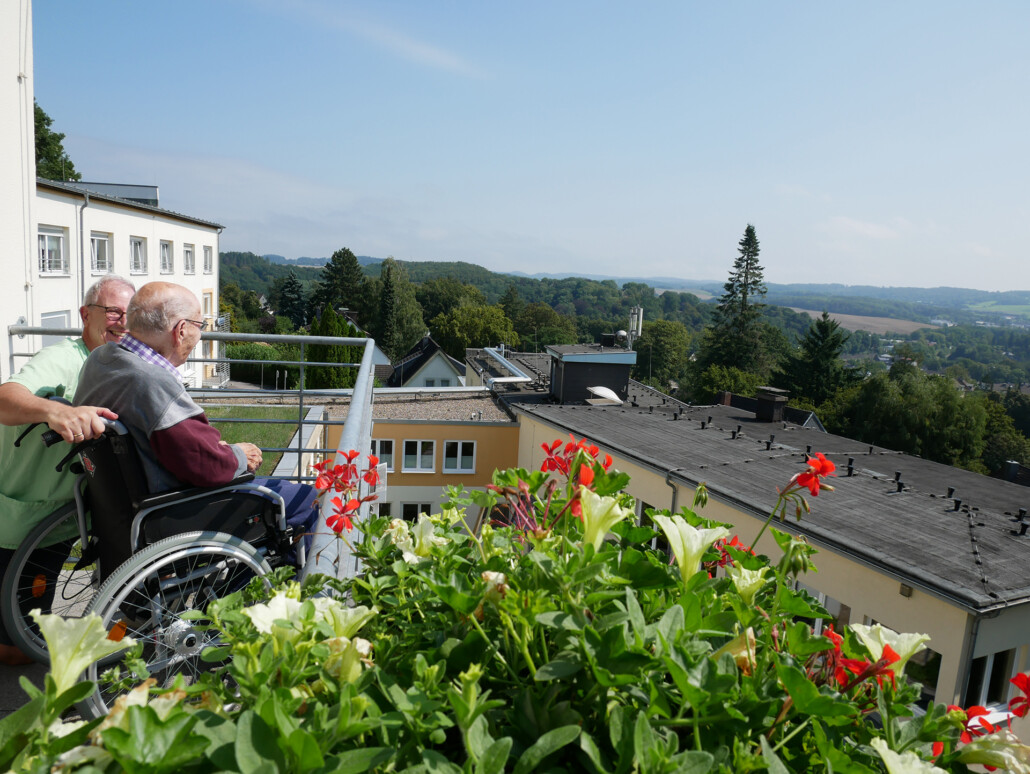 Pflegekraft begleitet einen Senior im Rollstuhl auf den Balkon, beide genießen den weiten Ausblick über die grüne Landschaft.